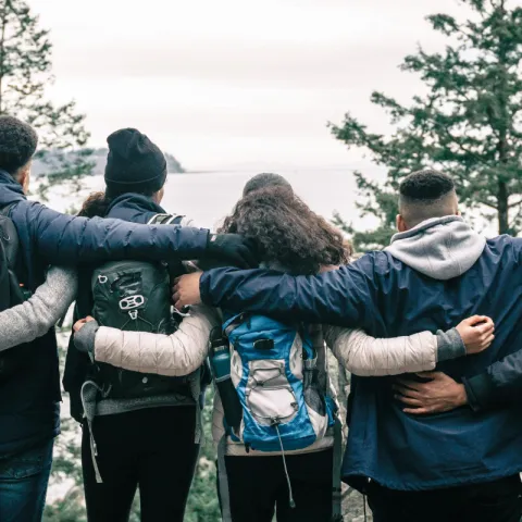 A diverse group of people wearing jackets and backpacks stand side by side with their arms around each other, facing a lake surrounded by evergreen trees.