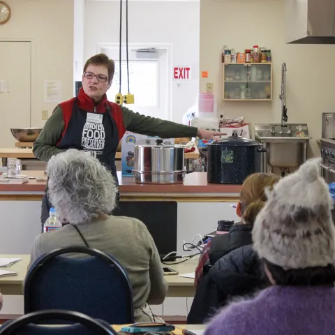 A woman standing in a commercial kitchen in front of an audience teaching.