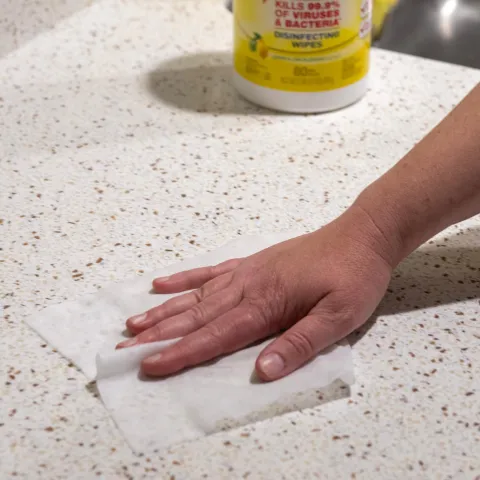 A person's arm wiping a white marble kitchen countertop with a white disinfectant wipe.