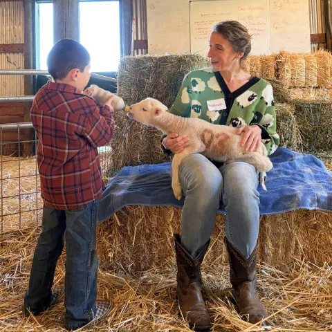 Community educator Hannah Bird holds a lamb on her lap as a schoolchild feeds it a bottle in a barn-like setting
