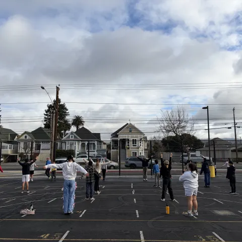 A group of elementary school children in a PE class outside on the school blacktop with their PE teacher on a cloudy day.