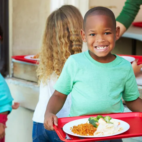 A young boy holds a red tray with a plate of fresh, school food while standing in line at the school cafeteria.
