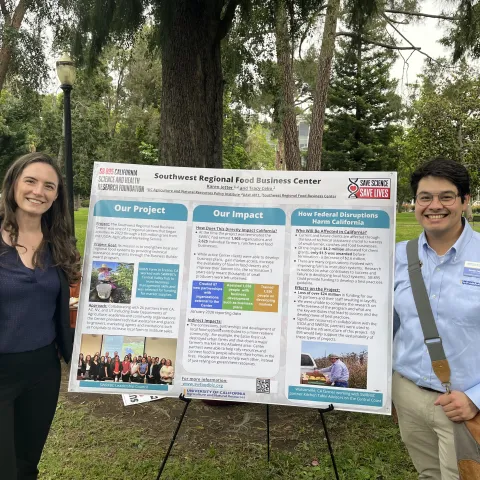 Anne and Nestor stand on opposite sides of a Southwest Regional Food Business Center poster describing the project, its impact and how federal disruptions harm California.