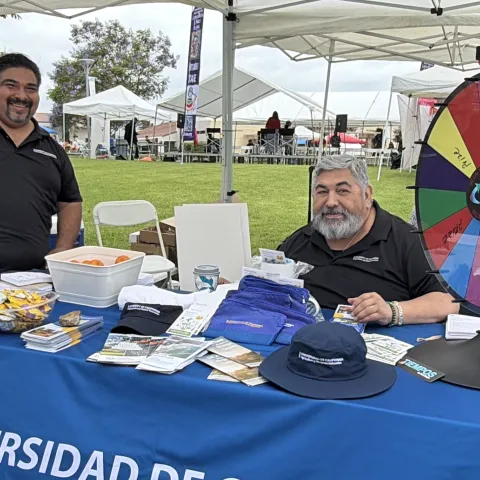 Miguel Sánchez y Ricardo Vela en una mesa de promoción de UC ANR, una ruleta y artículos de promoción sobre una mesa.
