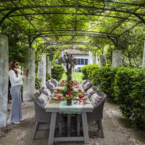 A long outdoor dining table is set with bright plates and linens underneath a lush, vine-covered metal pergola. The structure has thick weathered stone pillar legs. A woman in a white jacket and grey wide-leg trousers stands near a pillar, holding her phone to take a photo. Grey wicker armchairs with floral patterned cushions flank the table. The path is paved with large rectangular stones and box hedges, vining plants, and trees surround the area.