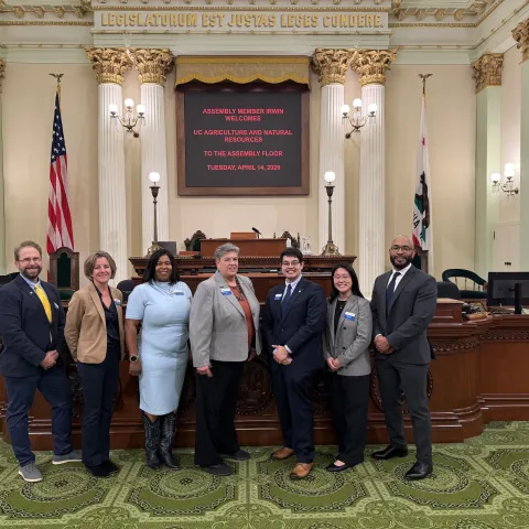 Seven people stand in a line in ornate chamber with pillars. Signs read: Legislatorum est justas leges condere. Assemblymember Irwin welcomes UC Agriculture and natural resources to the Assembly floor Tuesday, April 14, 2026
