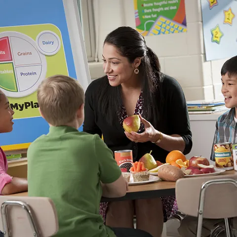 A teacher sits at a table with elementary-school aged students and talks with them about healthy eating, while holding a piece of fruit in her hands.