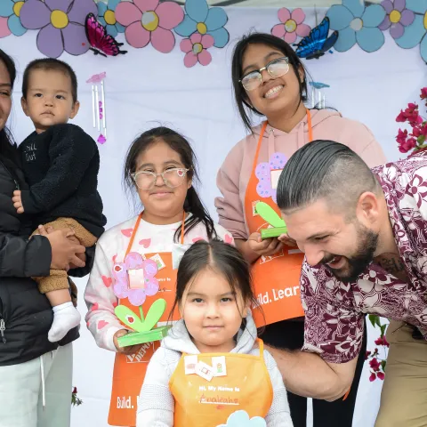 A family smiles while standing in a booth with artistic colors in the background. The family of six shows three of the four children wearing Home Depot orange aprons and holding floral crafts.