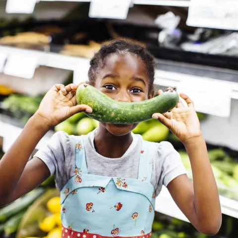 A young girl holds a cucumber in front of her face while standing in a grocery store produce aisle.