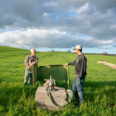 Two farmers stand in a vast grazing field, looking at a well pump