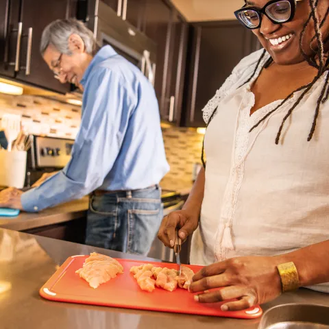 A woman and her husband stand in the kitchen while preparing a meal, she is cutting raw chicken on an orange cutting board.