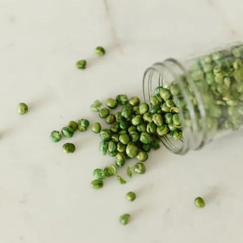 High-angle view of green peas spilling out of a glass jar onto a white surface.