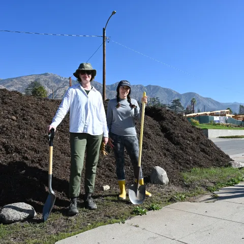 Two people with shovels stand in front of a pile of compost.