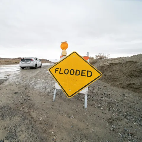 A road sign that reads "Flooded" in front of a car that is on a flooded street