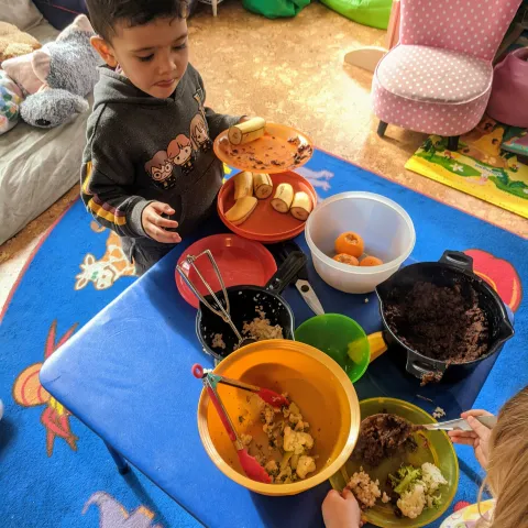 Two young children serve themselves food from colorful bowls set atop a low-table in a family child care home setting.
