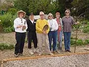 A group of six people poses in a community garden, holding gardening tools and wearing casual clothing, with garden beds visible in the background.