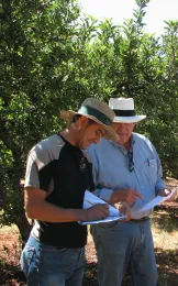 Two men under apple trees looking at a clip board.