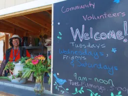 A volunteer stands behind a flower-filled counter at a farm stand, with a chalkboard sign displaying welcoming text and operational hours.