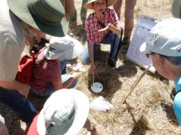 A female instructor leads a soil class outdoors, demonstrating soil testing techniques to engaged participants seated around her in a grassy area.