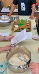 Table top surrounded my master food preservers. Table filled with items like a cooking pot, cutting board, jar, wooden spoon, and thermometer.