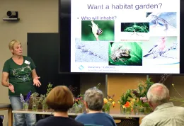 A Master Gardener presents on habitat gardens, gesturing to a slide featuring images of various wildlife. Attendees are seated, with jars of flowers on the table in front of them.
