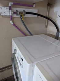 A washing machine beside a graywater system featuring PVC pipes labeled "To Sewer" and "To Landscape," with a yellow valve and black hose mounted on the wall.