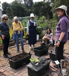Group of participants at a gardening workshop, engaging around planting boxes in a garden, with one woman leading the discussion.