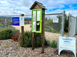 A bright green Seed Share structure with a glass panel and slanted roof in a garden setting. Surrounding signs indicate garden hours and details, with wood chips covering the ground.