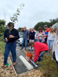 Jim holds an oak sapling in a container as a group of people in a parking lot observe.