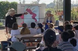 Two women present to a group of children outdoors, with an illustration of a pink figure on a whiteboard.
If you have specific details from the image, feel free to share them, and I can help refine the description further!