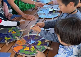 Children at a table are collaborating on an activity illustrating flower parts, using colored wedges of paper and labeled clothespins attached to a pie-shaped plate.