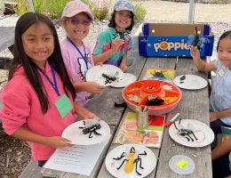 Four children at a picnic table each hold a plate with handmade bug creations made from crafting supplies in an orange container.