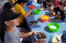 A group of school children sits at a table with egg carton containers for planting seeds, surrounded by colorful bowls filled with markers and planting supplies.