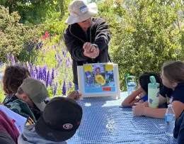 A Master Gardener instructs a group of children at a table outdoors, holding a display of yellow and purple flowers.