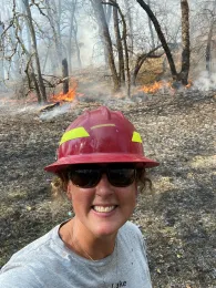 Lenya Quinn-Davidson is wearing a red hard hat with a prescribed fire buring in the background in an oak woodland in the winter
