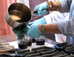 Gloved hands pouring elderberry syrup from a saucepan into a jar.