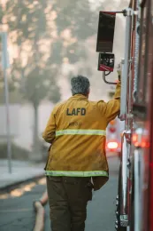 Firefighter leans against fire engine. LAFD printed on back of coat.