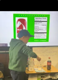 Child measuring teaspoons of sugar with a bottle of soda next to him