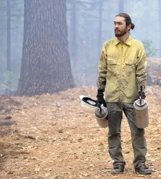Kane, wearing a yellow long-sleeved shirt, holds a hard hat and 2 drip torches in a forest 