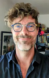 Jadrian Johnson smiles while wearing dark-rimmed glasses and a dark-colored button up shirt.