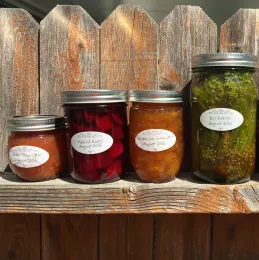 4 different sized jars containing beets, pickles, and jam lined up on a fence. 