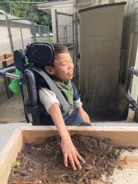 Young boy seated and strapped into a wheelchair seated adjacent to an elevated garden box, touching dirt with his left hand and smiling with joy