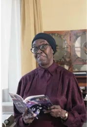 Gwendolyn Brooks sitting in a room holding a book wearing a red shirt, glasses and a blue headscarf