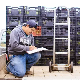 farmer kneeling next to pallets of produce, looking at clipboard