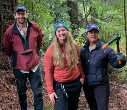 three participants in a forestry workshop stand in a forest with a tool