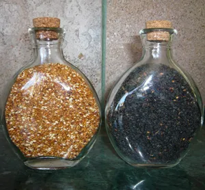 Two glass containers filled with toasted sesame seeds: one with golden-brown seeds and the other with dark black seeds, both sealed with cork stoppers.