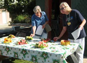 Two women participate in a watermelon tasting event outdoors, standing by a table filled with assorted watermelon pieces on a palm-patterned tablecloth.