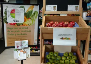 Baskets of apples in a small market are displayed along with Spanish and English language signs encouraging customers to eat fruits and vegetables
