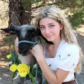 Blonde-haired girl poses next to lamb that is wearing a wreath of yellow roses around its neck.