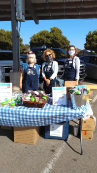 Three people with blue aprons in a parking lot standing in front of a table with information and materials